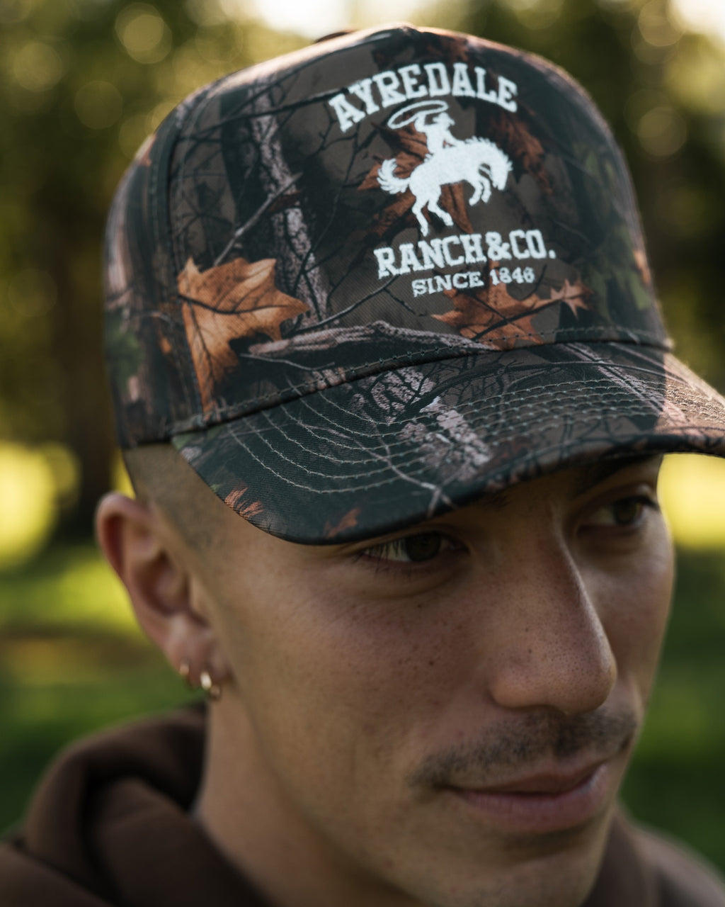 Ayredale Ranch & Co. camouflage baseball cap with white embroidered logo of a cowboy on a horse.