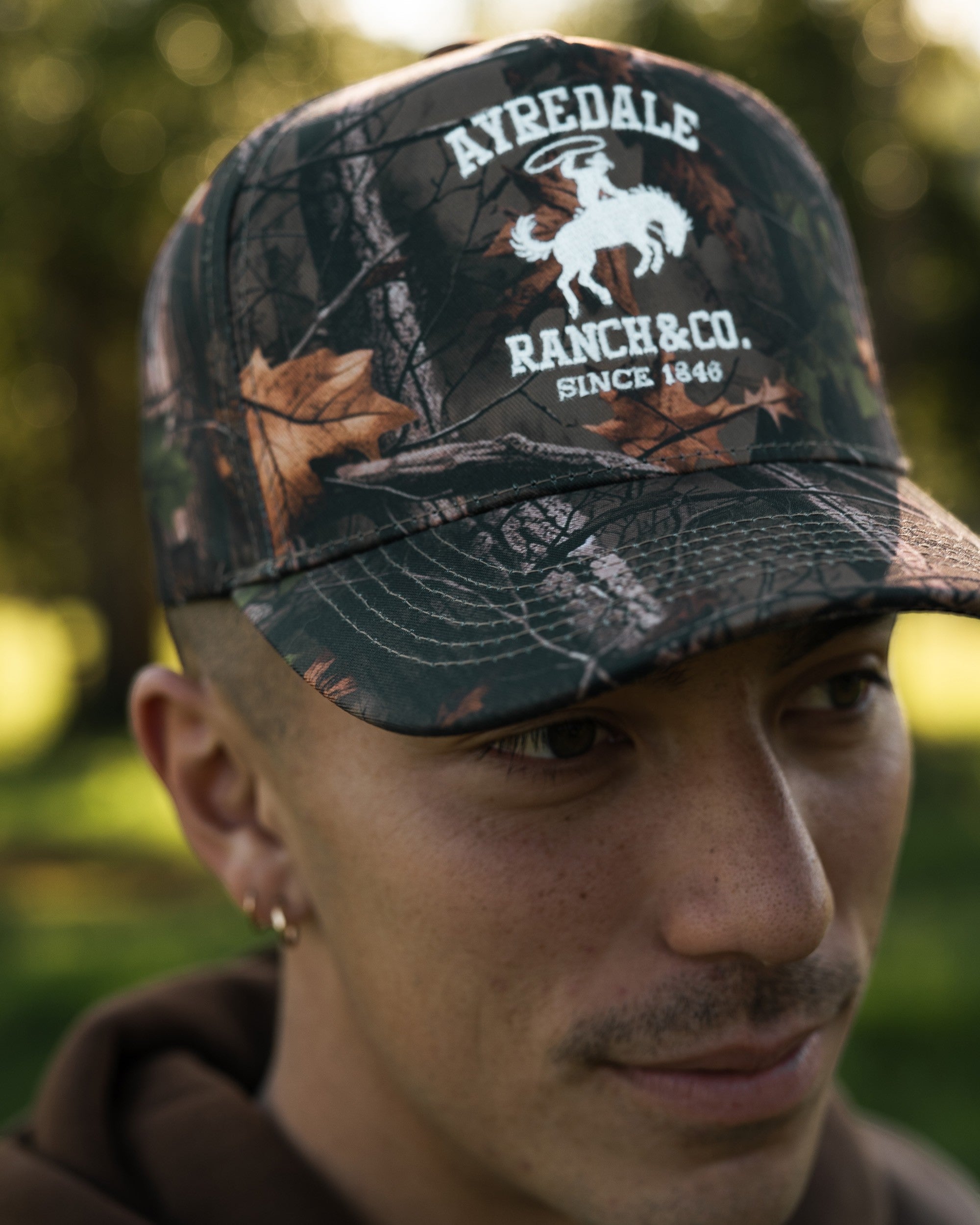Ayredale Ranch & Co. camouflage baseball cap with white embroidered logo of a cowboy on a horse.