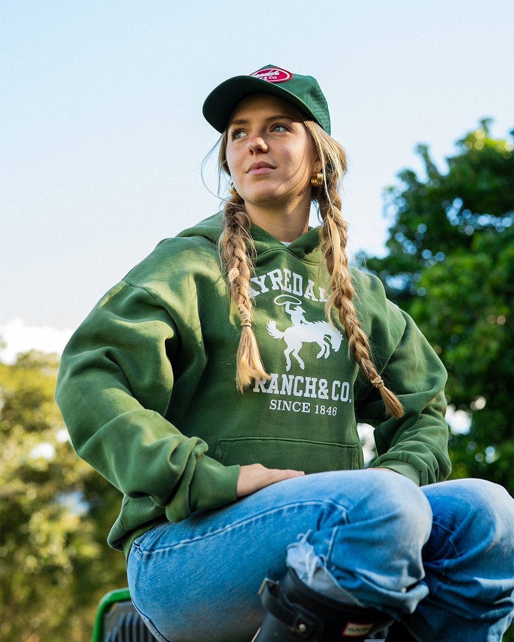 Woman wearing an Ayredale Ranch & Co. green heavyweight fleece hoodie with a collegiate graphic and matching cap