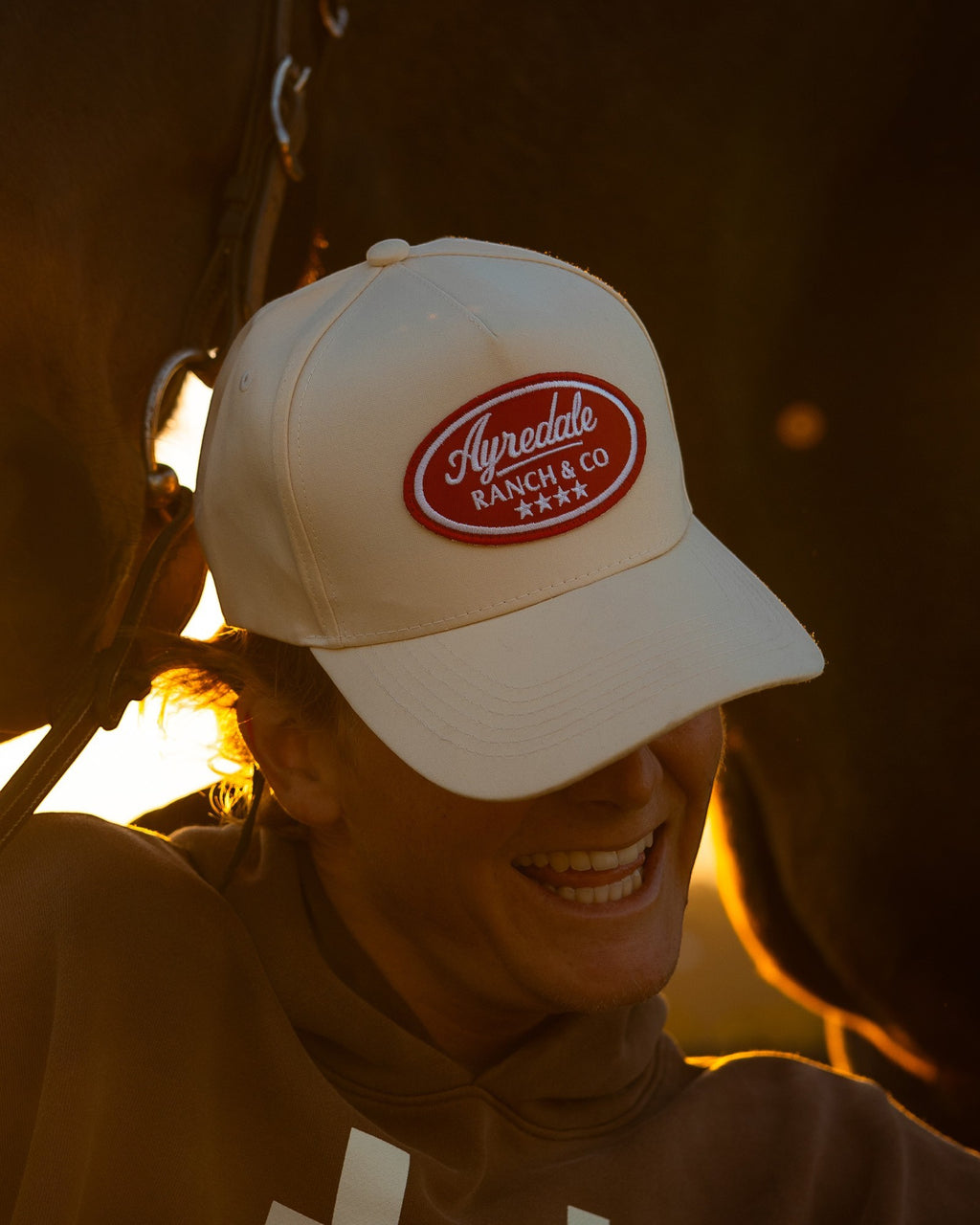 Ayredale Ranch & Co. white cotton-twill snapback hat with a red oval patch, worn by a smiling person at sunset.